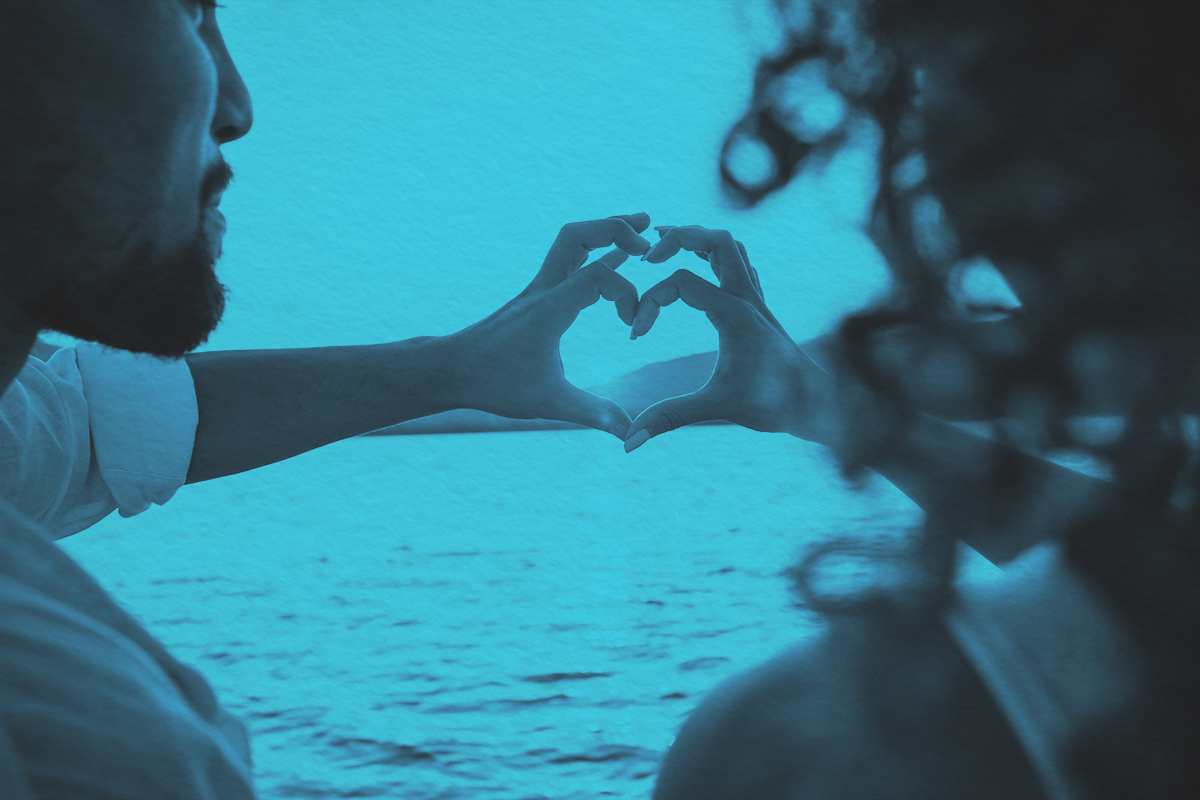 A couple doing heart shape with fingers looking at the sun setting behind the mountains