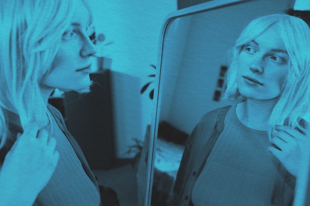 Portrait of young adult woman, standing in front of mirror touching hair and examining appearance